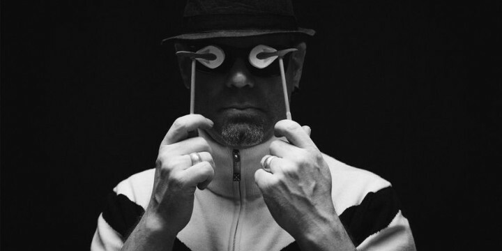 Schwarz-weiß Foto eines Mannes mit Hut, der sich an Stäbchen befindliche Objekte vor die Augen hält