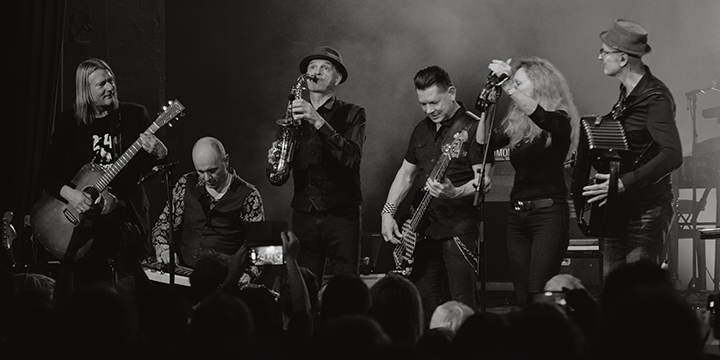 Schwarz-Weiß-Foto von einer 6-köpfigen Band, die Gitarre, Saxophon, Ziehharmonika spielen und die Frau singt.