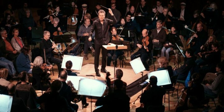 Dirigent Simon Gaudenz steht in der Mitte, um ihn herum kreisförmig Musiker:innen der Jenaer Philharmonie und dazwischen Publikum