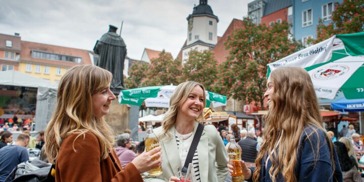 3 Mädels stehen mit Getränken auf dem Marktplatz vor einer Bühne und Biertischgarnituren, eingebettet vom historischen Marktlatz in Jena.  ©JenaKultur, R. Möbius