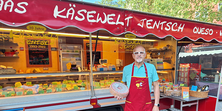 Ein Mann mit Käselaib im Arm vor einem mobilen Stand mit Käsespezialitäten in der Auslage auf dem Jenaer Wochenmarkt