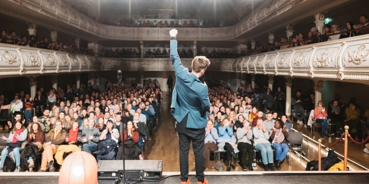 Comedian Friedrich Herrmann steht in einem prunkvollen Saal auf der Bühne und reißt seinen Arm in die Höhe, das Publikum applaudiert begeistert
