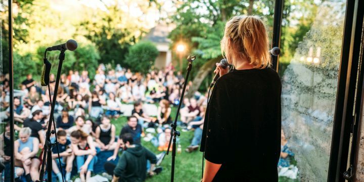 Publikum sitzt in einem Garten, im Vordergrund spricht eine junge Frau zu ihnen in ein Mikrofon