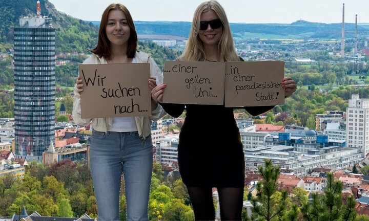 Zwei junge Frauen stehen vor einem Hintergrund mit Blick auf Jena und halten Schilder, auf denen steht, dass sie nach einer geilen Uni in einer paradiesischen Stadt suchen