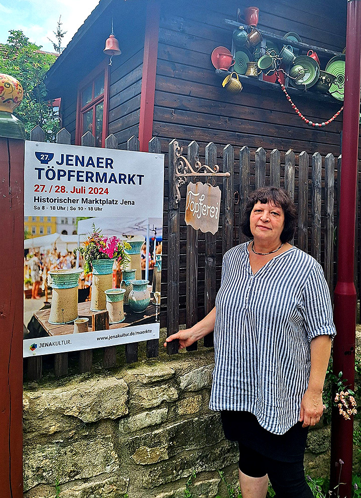 Töpferin vor einer Holzhütte mit Keramikobjekten und einem Plakat für den Jenaer Töpfermarkt