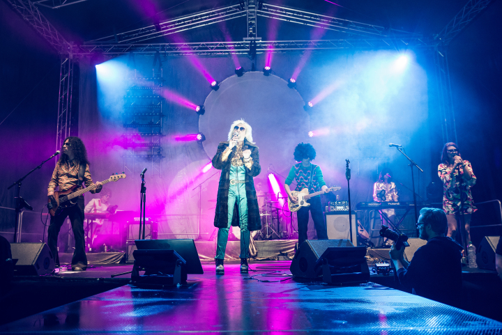 Band im Stile der 70er stehen auf einer Bühne und performen mit Gesang, Keyboard, Gitarre