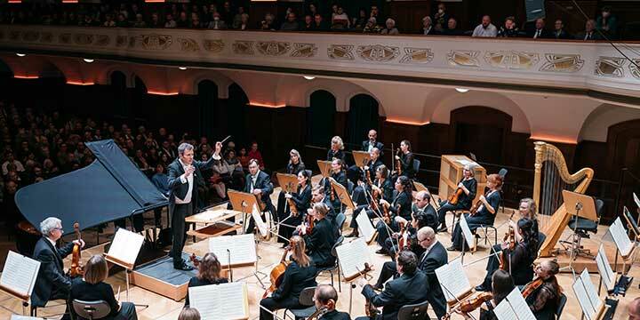 Orchestermusiker*innen der Jenaer Philharmonie in schwarzen Anzügen auf einer Bühnen. Der Dirigent steht vor den Musikerinnen mit gehoben Taktstock. Im Hintergrund in das Publikum auf Parkett und Loge.  ©JenaKultur, C. Worsch