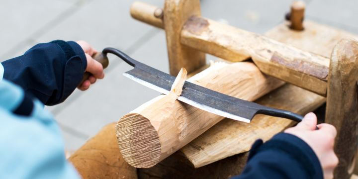 Kinderhände führen ein Holzbearbeitungswerkzeug mit Klinge über ein eingespanntes Stück Holz