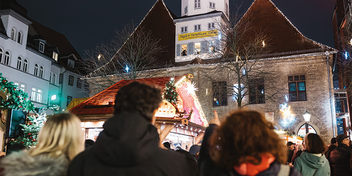 3 Menschen auf dem Jenaer Weihnachtsmarkt stehen mit dem Rücken zur Kamera und zeigen auf die Bläser im Rathausturm